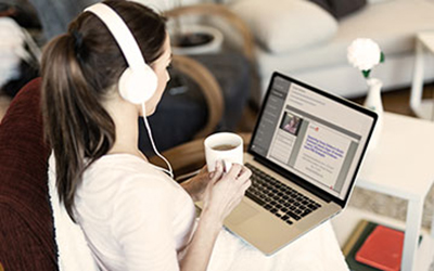 woman in headphones drinking coffee in her living room while she watches a live webinar on her laptop